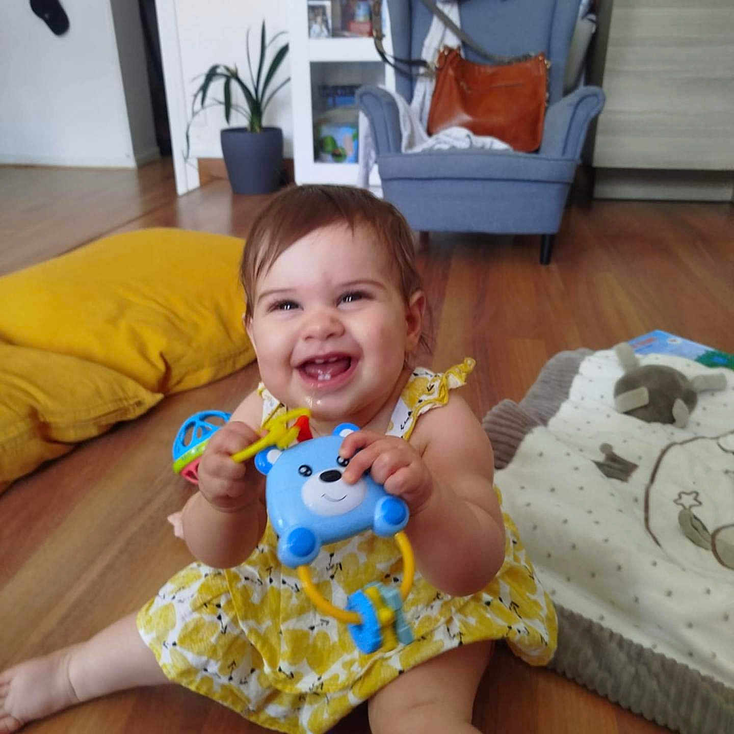 Emma a rejoint le concours — aidez-le/la à gagner de superbes lots ! armchair, baby, cheerful, child, cushion, cute, face, floor, happy, indoor, infant, living_room, pacifier, person, play, potted_plant, smile, toy, wooden_floor, yellow_dress