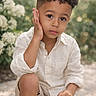 child, boy, curly_hair, outdoor, nature, greenery, flower, portrait, casual_clothing, white_shirt, shorts, sitting, thoughtful, young, daylight, skin, face, hand, natural_light, expression