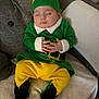 baby, sleeping, elf_costume, green_clothing, yellow_pants, hat, couch, blanket, infant, hands, closed_eyes, indoor, soft_texture, cute, holiday, festive, child, portrait, relaxation, cozy