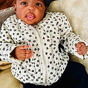 Na'Kyah is registered to the contest to win money with this photo: baby, child, black_bow, jacket, flower_pattern, tongue_out, cozy, blanket, indoor, couch, cute, infant, portrait, smiling, happy, young_child, clothing, socks, relaxed, wooden_floor