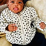 baby, child, black_bow, jacket, flower_pattern, tongue_out, cozy, blanket, indoor, couch, cute, infant, portrait, smiling, happy, young_child, clothing, socks, relaxed, wooden_floor