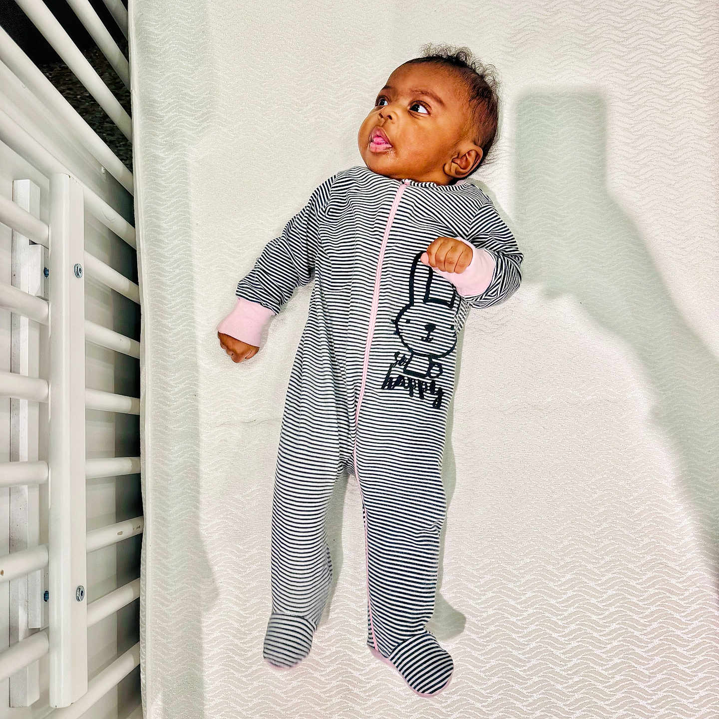 Na'Kyah is registered to the contest to win money with this photo: baby, baby_clothes, bunny_print, child, crib, curious, cute, footed_pajamas, indoor, infant, lying_down, mattress, newborn, onesie, person, portrait, shadow, sleepwear, striped_clothing, white