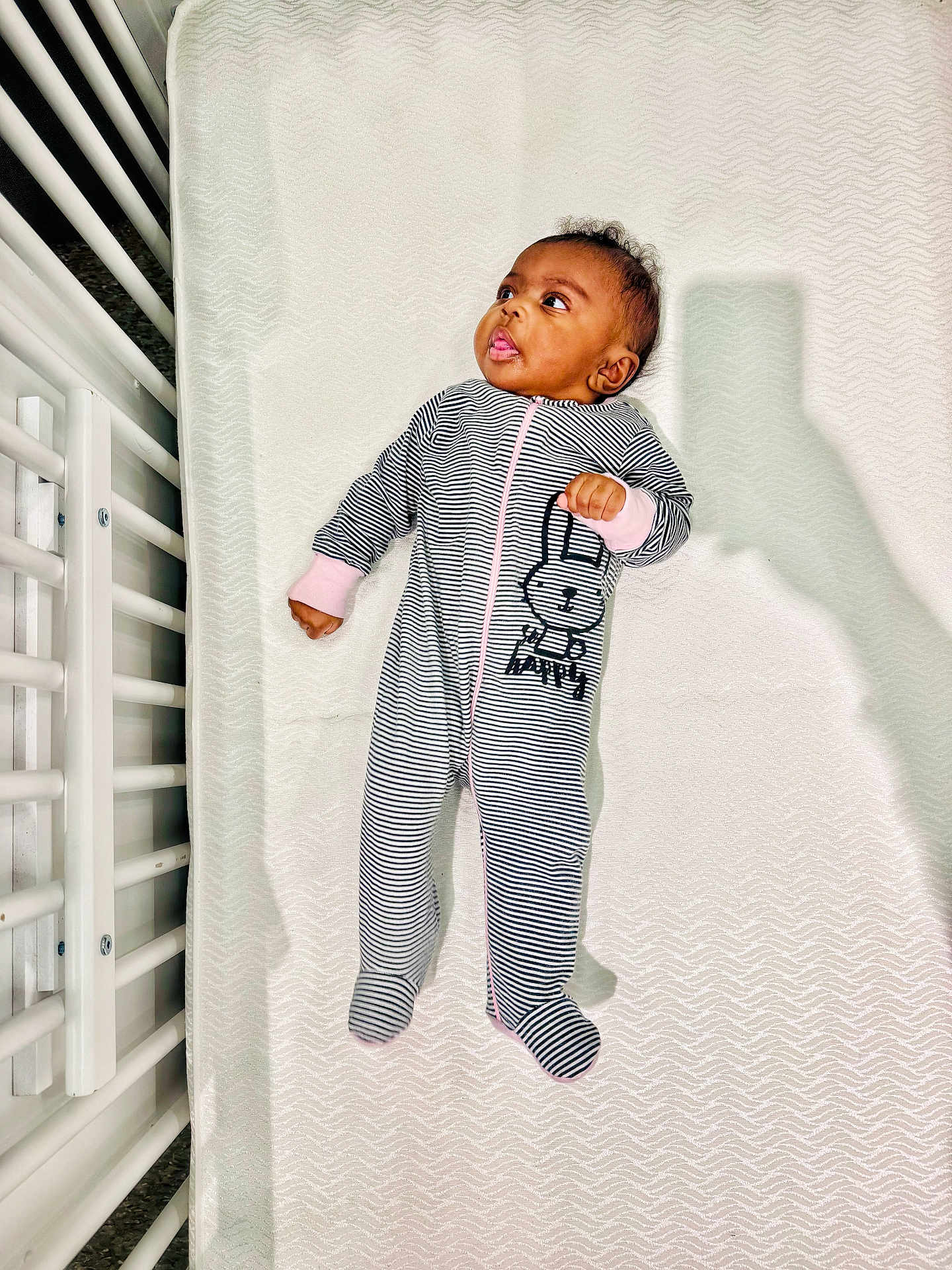 Na'Kyah is registered to the contest to win money with this photo: baby, infant, onesie, striped_clothing, bunny_print, crib, mattress, white, shadow, curious, lying_down, baby_clothes, footed_pajamas, portrait, indoor, child, person, sleepwear, cute, newborn