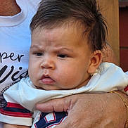 Nickolas is registered to the contest to win money with this photo: baby, child, infant, face, expression, furrowed_brow, hair, hand, adult_hand, clothing, shirt, white_shirt, red_stripe, blue_stripe, holding, portrait, closeup, skin, person, outdoor
