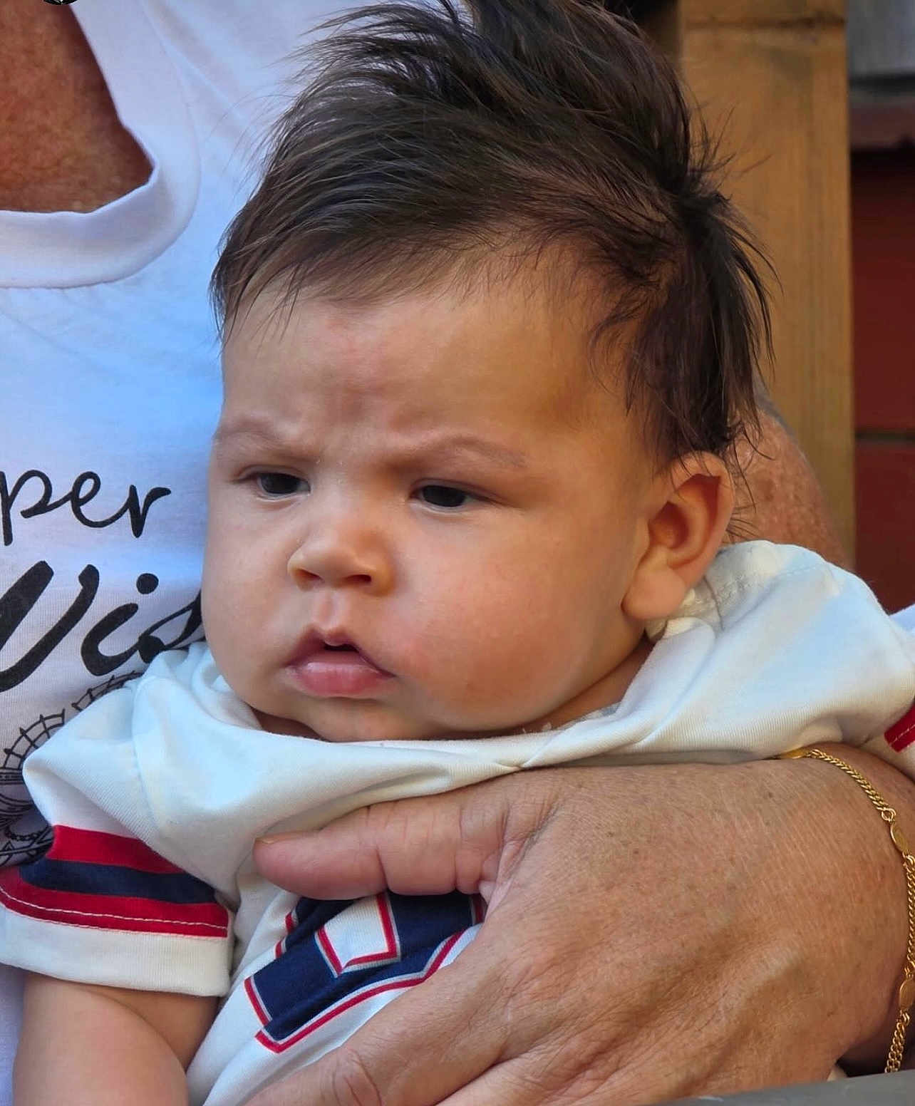 Nickolas is registered to the contest to win money with this photo: baby, child, infant, face, expression, furrowed_brow, hair, hand, adult_hand, clothing, shirt, white_shirt, red_stripe, blue_stripe, holding, portrait, closeup, skin, person, outdoor