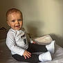 black_pants, bright_eyes, cartoon_print, casual_clothing, child, goose_pattern, happy, indoor, joyful, light_skin, neutral_background, padded_surface, portrait, short_hair, sitting, smiling, toddler, white_shirt, white_sneakers, young_child