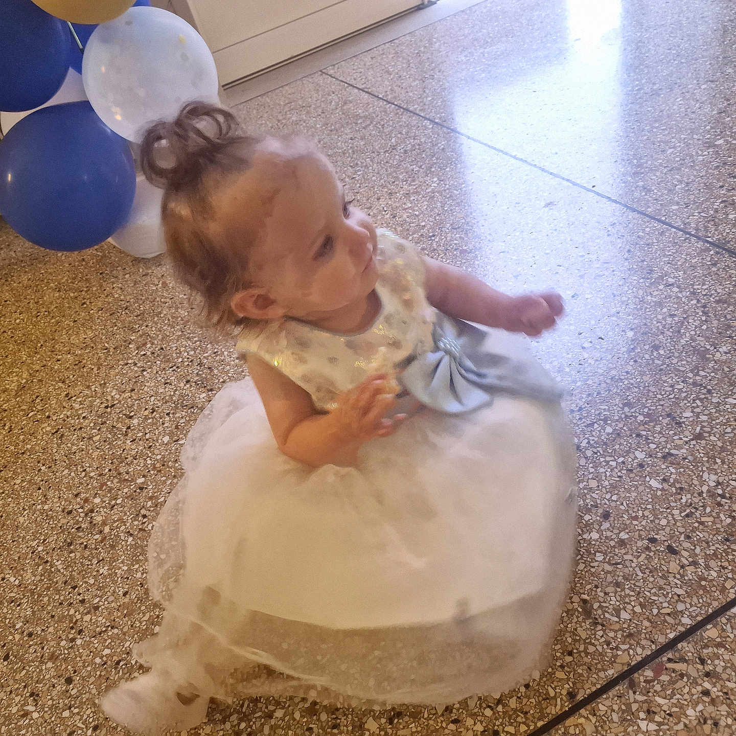 Luna participe au concours pour gagner de l'argent avec cette photo : baby, balloons, bow, celebration, child, curly_hair, cute, dress, festive, floor, floor_tiles, happy, indoor, looking_away, party, person, sitting, small_child, toddler, white_dress