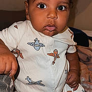 Jaden is registered to the contest to win money with this photo: baby, child, infant, curly_hair, onesie, airplane_pattern, drool, indoor, blanket, soft_lighting, closeup, portrait, cute, wide_eyes, seated, clothing, hand, skin, expression, young