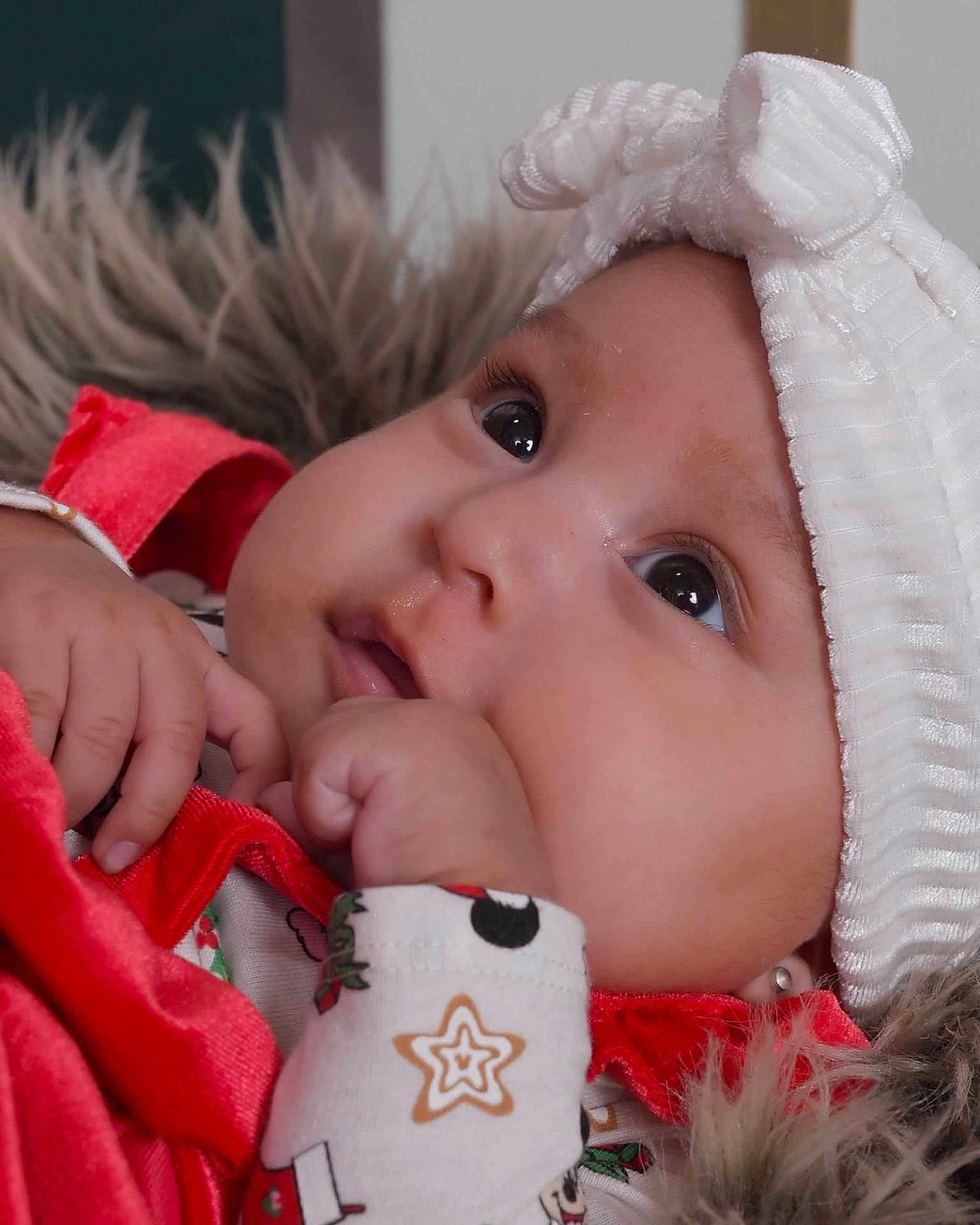 Veronica is registered to the contest to win money with this photo: baby, infant, close_up, headband, bow, red_clothing, festive, hand, face, eyes, skin, fur_background, cute, adorable, portrait, soft_texture, young_child, sleepwear, detail, expression