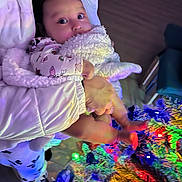 Veronica joined the competition — help win amazing prizes! baby, blanket, child, christmas_tree, colorful_lights, decorations, indoors, person, holiday, festive, holding, floor, wooden_floor, winter_clothing, cute, infant, hand, feet, warm, expression