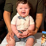 Kenai joined the competition — help win amazing prizes! baby, child, smiling, bow_tie, suspenders, table, adult_hands, green_shirt, happy, portrait, indoor, person, clothing, feet, hands, photograph, family, cute, face, infant