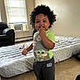 toddler, child, indoors, curly_hair, baby_yoda, shirt, pants, barefoot, bed, window, air_conditioner, floor, smiling, standing, person, living_room, blanket, furniture, happy, casual_clothing