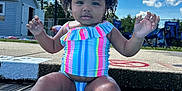 Harmonie joined the competition — help win amazing prizes! toddler, child, swimsuit, pool, water, outdoor, summer, sky, clouds, curly_hair, sitting, playful, happy, sunny, feet_in_water, concrete_edge, colorful_clothing, daytime, fence, plastic_chairs