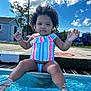 toddler, child, swimsuit, pool, water, outdoor, summer, sky, clouds, curly_hair, sitting, playful, happy, sunny, feet_in_water, concrete_edge, colorful_clothing, daytime, fence, plastic_chairs