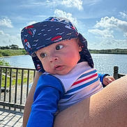 Alexys is registered to the contest to win money with this photo: baby, child, hat, blue_clothing, striped_shirt, outdoor, water, sky, clouds, fence, person, holding, portrait, sunhat, nature, daylight, cute, expression, infant, summer