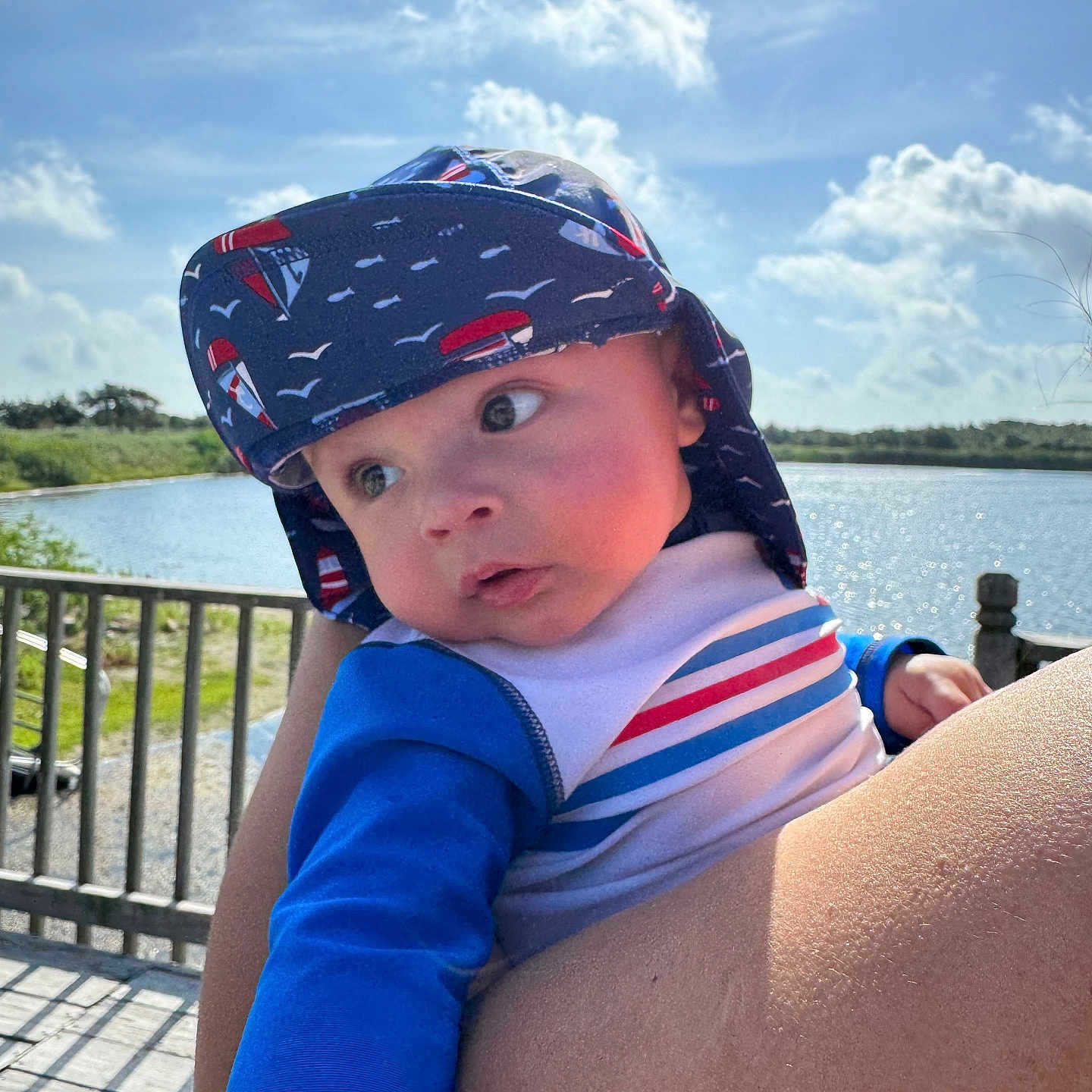Alexys is registered to the contest to win money with this photo: baby, blue_clothing, child, clouds, cute, daylight, expression, fence, hat, holding, infant, nature, outdoor, person, portrait, sky, striped_shirt, summer, sunhat, water