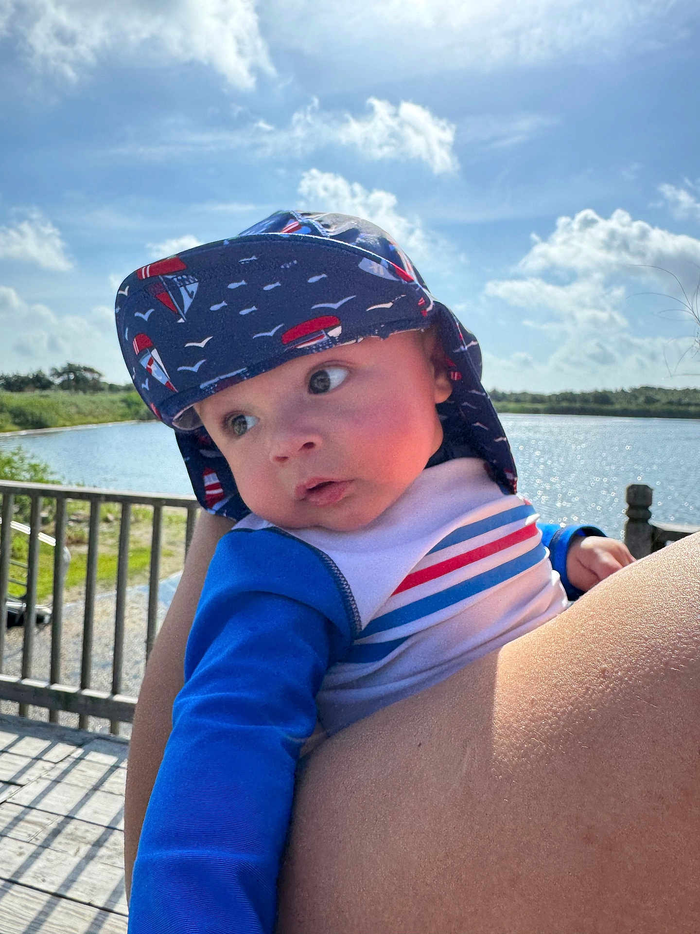 Alexys is registered to the contest to win money with this photo: baby, child, hat, blue_clothing, striped_shirt, outdoor, water, sky, clouds, fence, person, holding, portrait, sunhat, nature, daylight, cute, expression, infant, summer