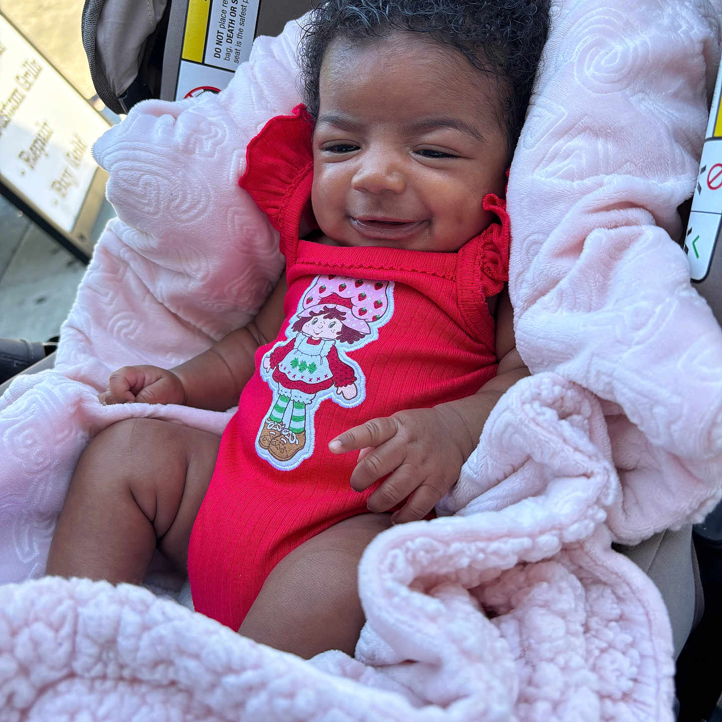 Melanii is registered to the contest to win money with this photo: baby, infant, smiling, blanket, car_seat, red_clothing, cute, happy, child, indoors, portrait, curly_hair, warning_label, soft_texture, seat, resting, cozy, small_hand, pink, cute_outfit