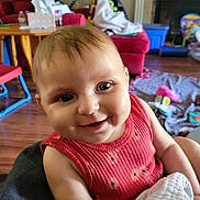 Clarity is registered to the contest to win money with this photo: arm, baby, blanket, child, clothing, face, floor, furniture, happy, head, home, indoor, infant, living_room, person, red_couch, seated, smiling, toy, wooden_table