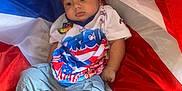 Kamari joined the competition — help win amazing prizes! baby, child, flag, puerto_rico, headband, shirt, pants, barefoot, fabric, red, blue, white, portrait, infant, clothing, person, sitting, indoors, cute, expression