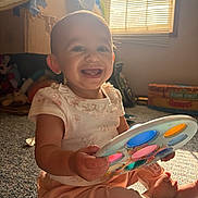Anya is registered to the contest to win money with this photo: baby, child, smiling, toy, indoor, carpet, window, sunlight, playful, happy, person, clothing, floor, colorful, face, sitting, infant, cute, light, home