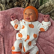 Davina is registered to the contest to win money with this photo: baby, infant, onesie, pumpkin_pattern, orange_hat, knitted_socks, jack_o_lantern, pink_blanket, soft_texture, cute, playful_expression, lying_down, indoors, newborn, clothing, face, hands, feet, smiling, child