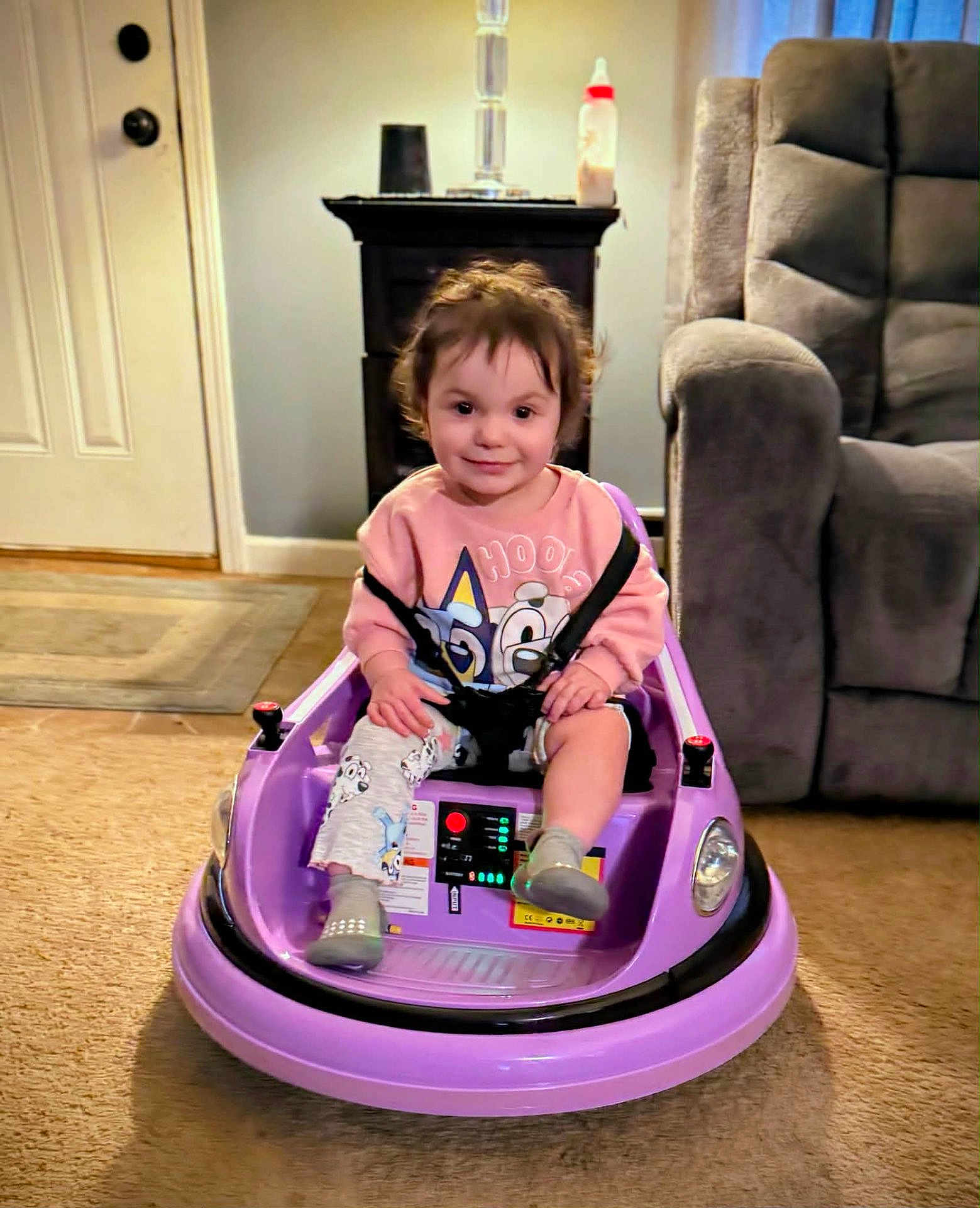Caralyn joined the competition — help win amazing prizes! child, toddler, toy_car, purple, smile, seatbelt, living_room, armchair, lamp, baby_bottle, carpet, floor, person, indoors, cute, hair, sweatshirt, playtime, furniture, portrait