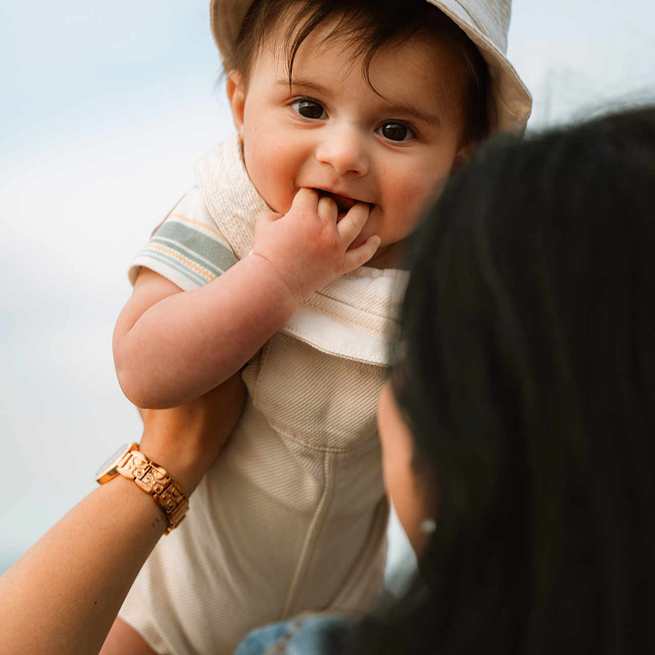 Vyom is registered to the contest to win money with this photo: adult_hand, baby, casual_clothing, child, cute, daylight, denim_jacket, face, fingers_in_mouth, happy, hat, holding, infant, outdoor, person, portrait, sky, smiling, watch, young_child
