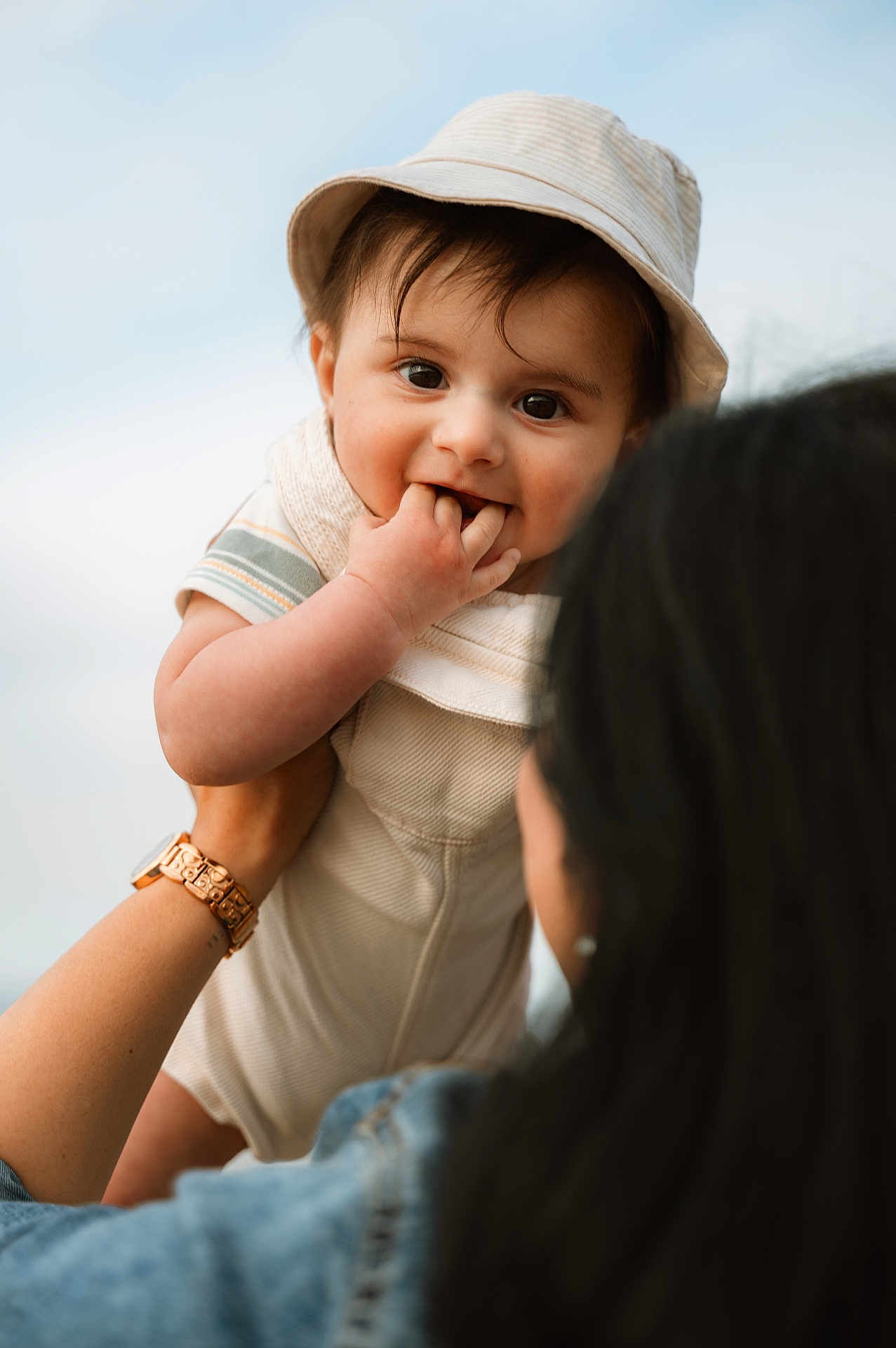 Vyom is registered to the contest to win money with this photo: baby, child, hat, smiling, fingers_in_mouth, adult_hand, watch, denim_jacket, outdoor, sky, portrait, cute, happy, infant, person, holding, face, young_child, casual_clothing, daylight