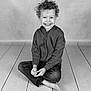 child, smiling, curly_hair, sitting, cross_legged, hoodie, jeans, barefoot, studio, black_and_white, floor, wooden_floor, portrait, happy, young, kid, casual_clothing, cute, innocence, joy