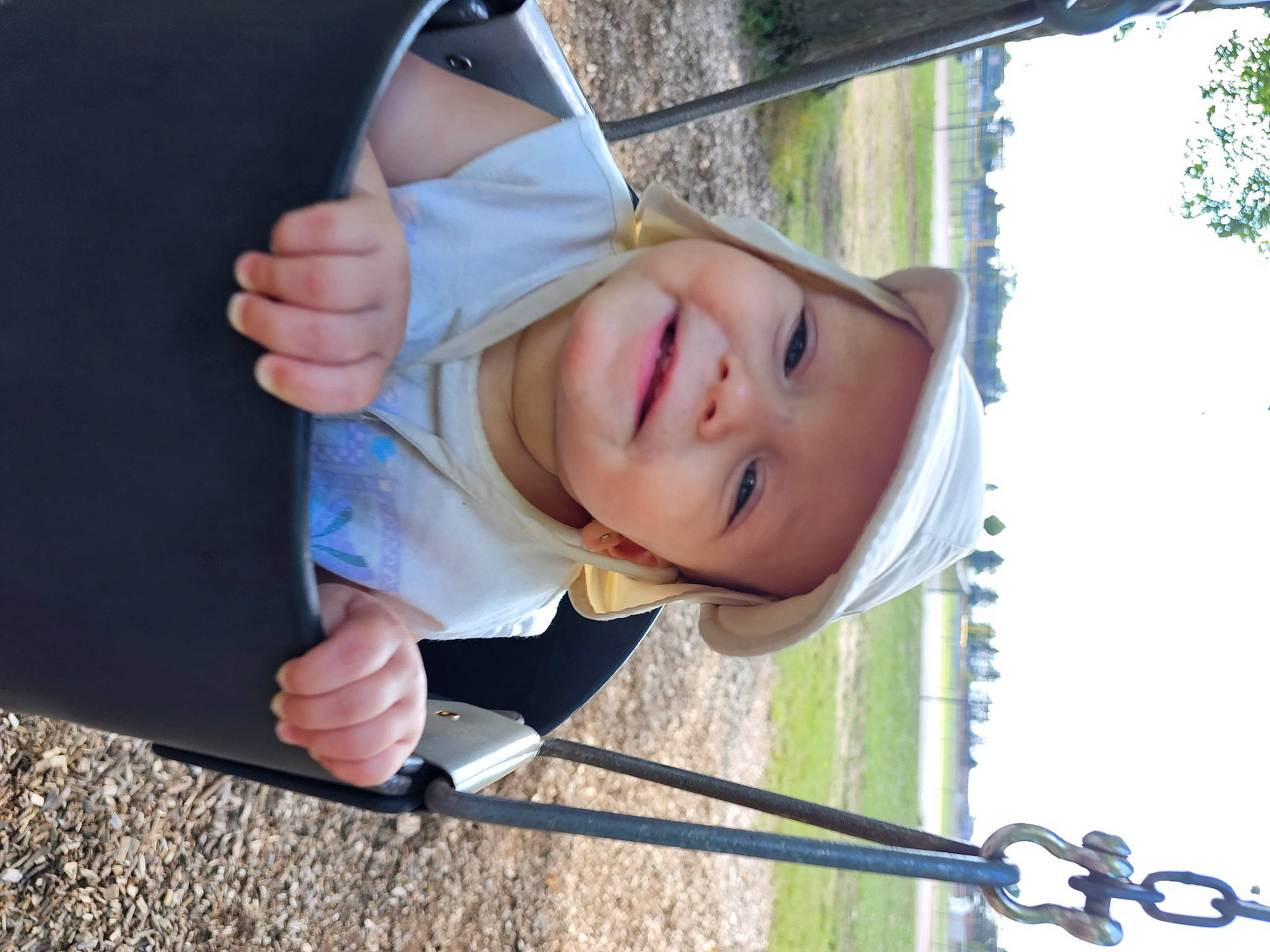 Maeve is registered to the contest to win money with this photo: child, city, fun, gesture, grass, happy, headwear, joy, leisure, outdoor_play_equipment, person, plant, play, playground, recreation, sky, sleeve, smile, swing, toddler