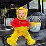baby, child, winnie_the_pooh, costume, yellow_outfit, red_shirt, pacifier, honey_pot, golf_cart, vehicle, seated, portrait, outdoor, sunlight, playful, costume_hat, safari, leather_seat, toy_bucket, cute