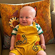 Harry is registered to the contest to win money with this photo: baby, bald_head, casual, child, colorful_clothing, comfortable, couch, cute, decorative_pillow, happy, home, indoor, infant, patterned_pillow, portrait, seated, smiling, soft_lighting, striped_shirt, yellow_overalls