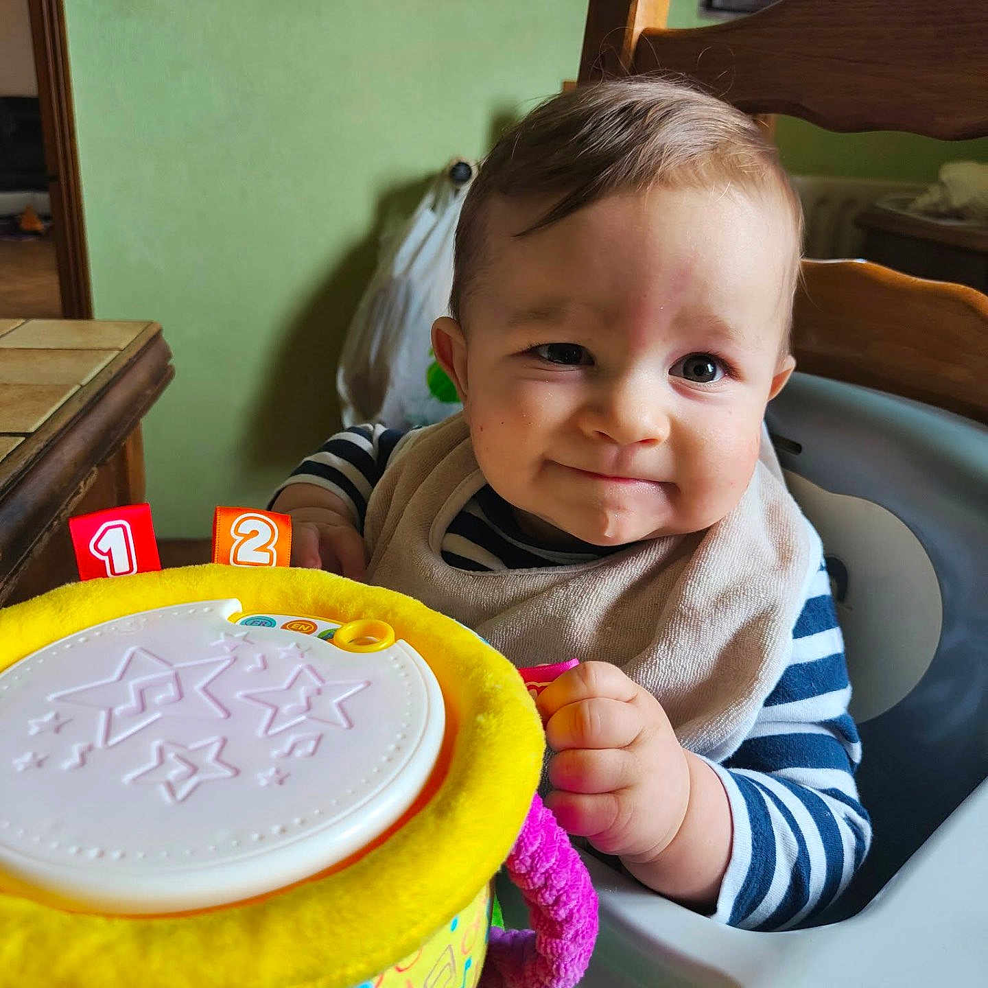 Elio a rejoint le concours — aidez-le/la à gagner de superbes lots ! baby, child, high_chair, toy, colorful, smile, face, indoor, clothing, striped_shirt, bib, hand, play, furniture, window, wood, table, green_wall, cute, person