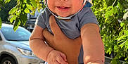 Noah joined the competition — help win amazing prizes! baby, child, person, hand, hat, outdoor, green_leaves, sunlight, car, sidewalk, smile, face, arm, clothing, shorts, holding, nature, daylight, happy, portrait
