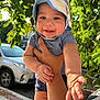 baby, child, person, hand, hat, outdoor, green_leaves, sunlight, car, sidewalk, smile, face, arm, clothing, shorts, holding, nature, daylight, happy, portrait