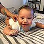 Noah joined the competition — help win amazing prizes! baby, smiling, bed, striped_blanket, hand, tattoo, person, indoor, happy, face, infant, clothing, sleeve, sitting, bedroom, shelf, furniture, home, cute, child