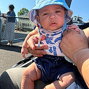 Noah joined the competition — help win amazing prizes! baby, child, hat, blue_hat, outdoor, sunny, person, holding, hand, clothing, shorts, infant, fence, background_people, casual, summer, seat, legs, expression, portrait