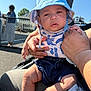 baby, child, hat, blue_hat, outdoor, sunny, person, holding, hand, clothing, shorts, infant, fence, background_people, casual, summer, seat, legs, expression, portrait