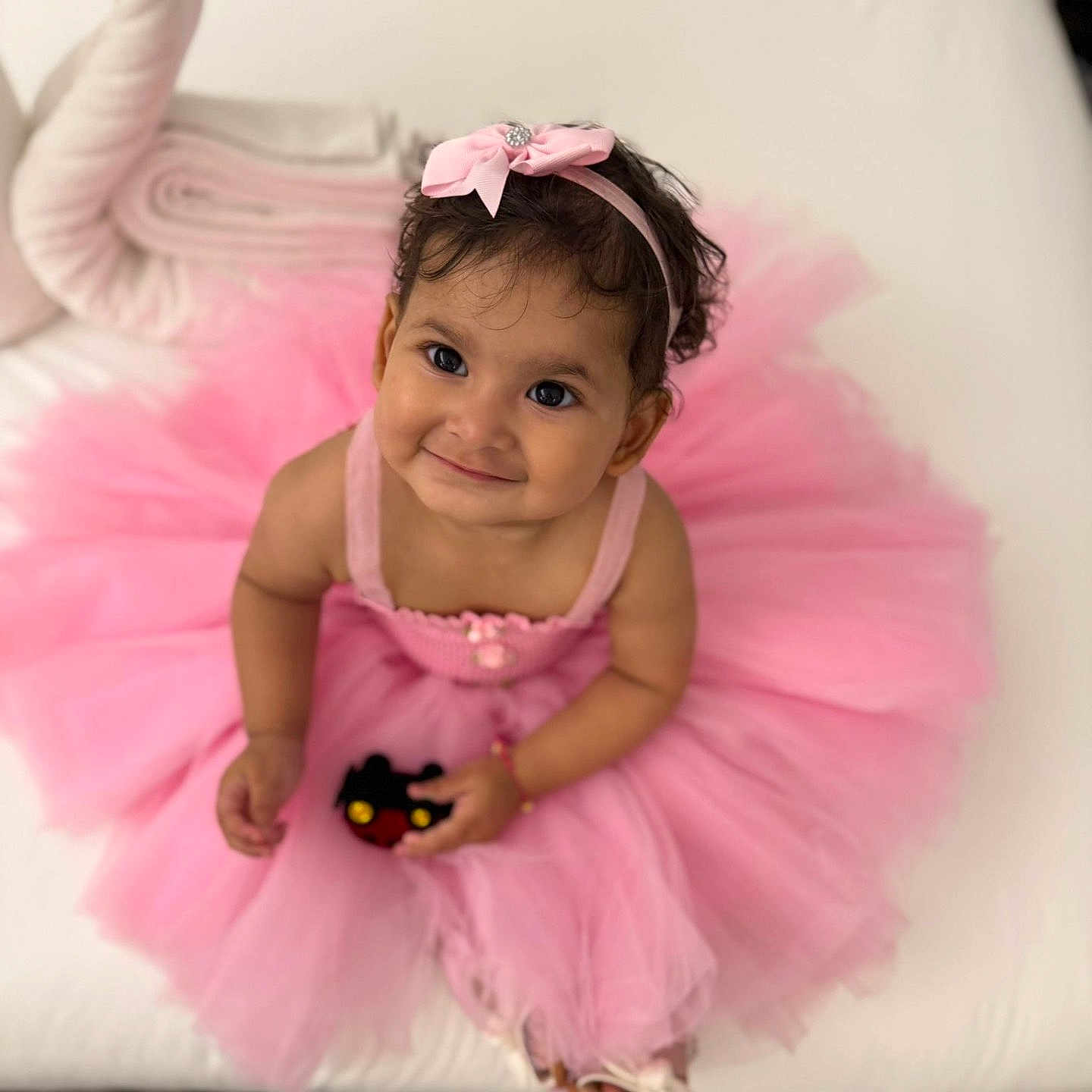 Smaya is registered to the contest to win money with this photo: accessory, baby, child, couch, curly_hair, cute, dress, footwear, girl, happy, headband, indoor, pink_tutu, playful, portrait, sitting, smiling, toddler, toy, young_child