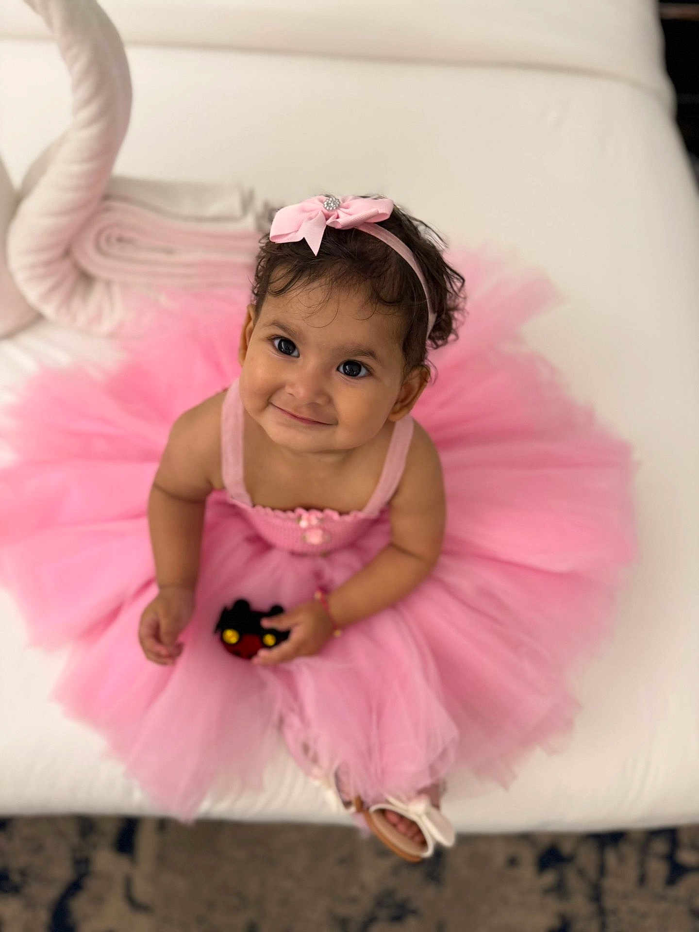Smaya is registered to the contest to win money with this photo: toddler, child, pink_tutu, headband, smiling, toy, indoor, couch, cute, portrait, dress, baby, sitting, footwear, happy, girl, curly_hair, playful, accessory, young_child