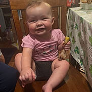 Autumn is registered to the contest to win money with this photo: baby, child, smiling, chair, wooden_chair, indoor, food, chicken_drumstick, table, tablecloth, shamrock_pattern, happy, person, clothing, pink_shirt, legs, hand, face, home, dining_area