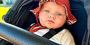 Bohdi is registered to the contest to win money with this photo: baby, stroller, hat, red_hat, child, face, blue_eyes, sunlight, outdoor, safety_harness, infant, clothing, seat, blurred_background, hand, expression, portrait, casual, daytime, person