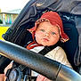baby, stroller, hat, red_hat, child, face, blue_eyes, sunlight, outdoor, safety_harness, infant, clothing, seat, blurred_background, hand, expression, portrait, casual, daytime, person
