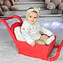 baby, child, red_sleigh, white_sweater, headband, beige_pants, teddy_bears, plush_toys, holiday_decor, festive_lights, indoor, portrait, smiling, cute, winter_clothing, carpet, cozy, decorative, childrens_photography, warm
