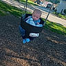 baby, swing, playground, child, outdoor, park, grass, trees, residential, daytime, blue_clothing, infant, seat, chain, woodchips, curious, person, sunlight, casual, expression
