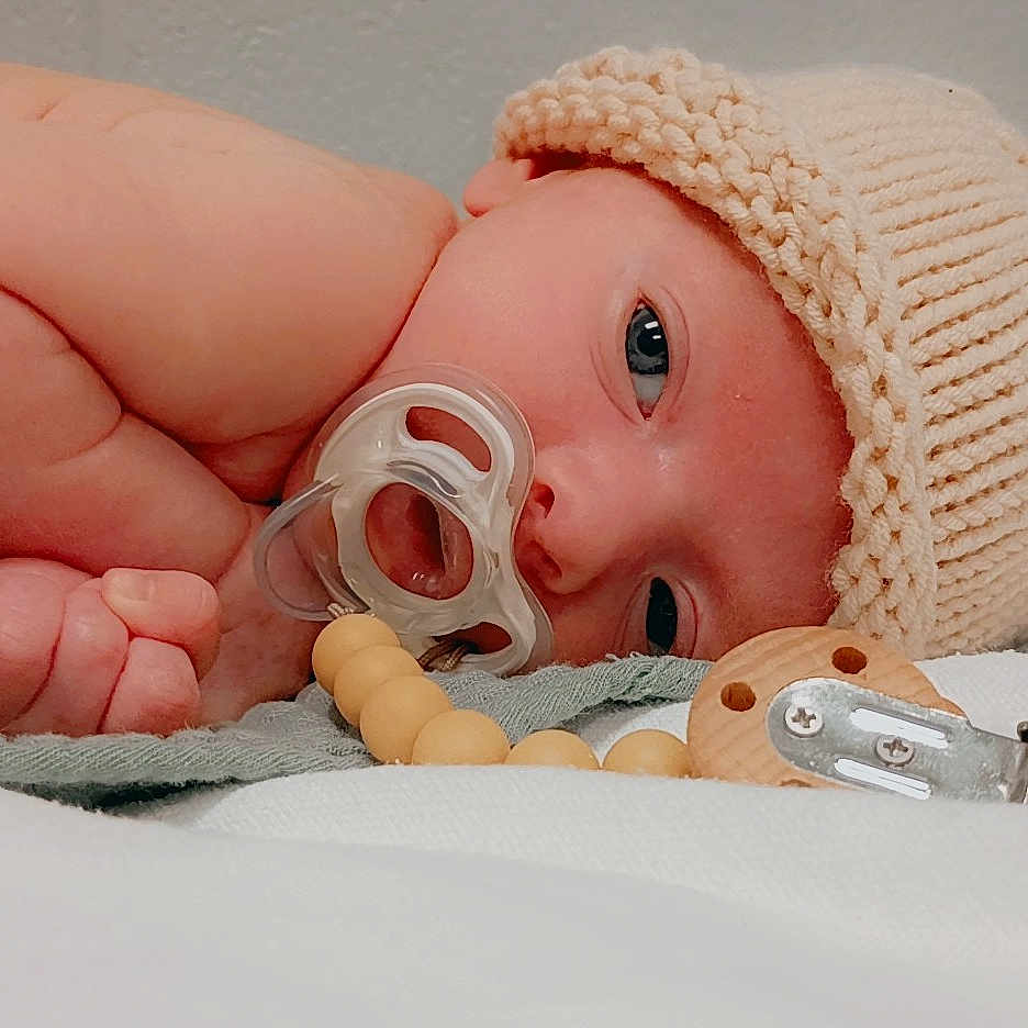 Harley is registered to the contest to win money with this photo: baby, infant, pacifier, knitted_hat, beads, lying_down, soft_surface, curious_eyes, hand, skin, cute, newborn, warm_colors, indoors, sleepwear, headwear, closeup, portrait, baby_accessory, resting