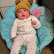 Angel is registered to the contest to win money with this photo: baby, infant, hat, pom_poms, yellow_hat, onesie, white_clothing, blue_chair, cushion, indoor, carpet, furniture, face, drool, cute, child, seated, expression, cozy, comfortable