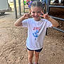 Lana is registered to the contest to win money with this photo: child, girl, smiling, peace_sign, hair_bows, tshirt, shorts, sneakers, dirt_ground, horse_stable, horse, person, outdoor, daylight, fence, rider, background, happy, casual_clothing, portrait