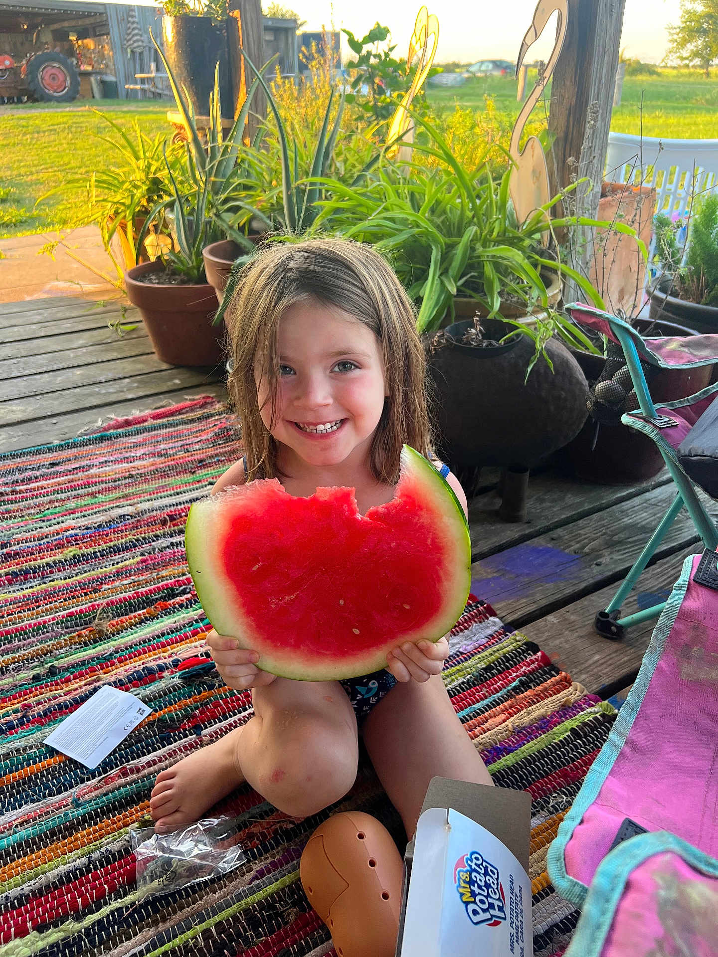 Lana is registered to the contest to win money with this photo: child, watermelon, smiling, outdoor, porch, plants, pottery, toy, mrs_potato_head, rug, wood, sunlight, happy, summer, playful, feet, clothing, nature, greenery, sitting